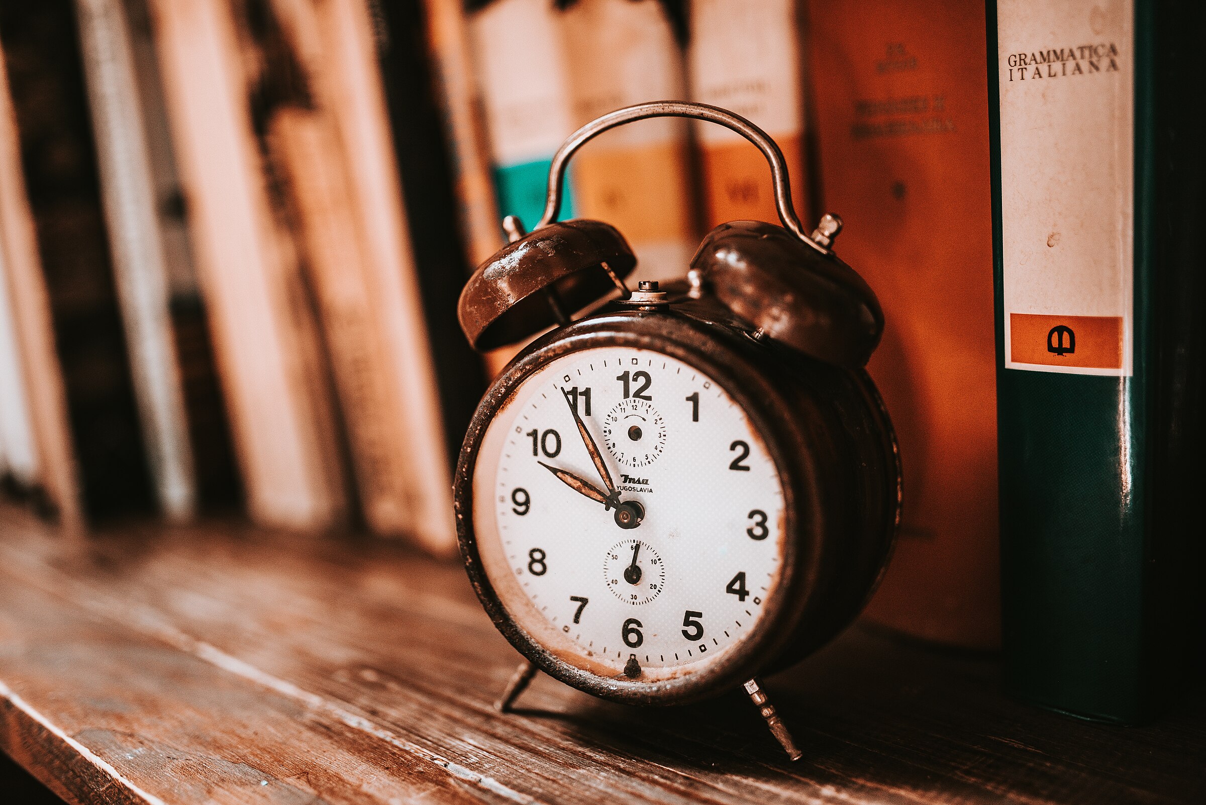 An old twin-bell wind-up alarm clock on a wooden shelf in front of a row of books.
