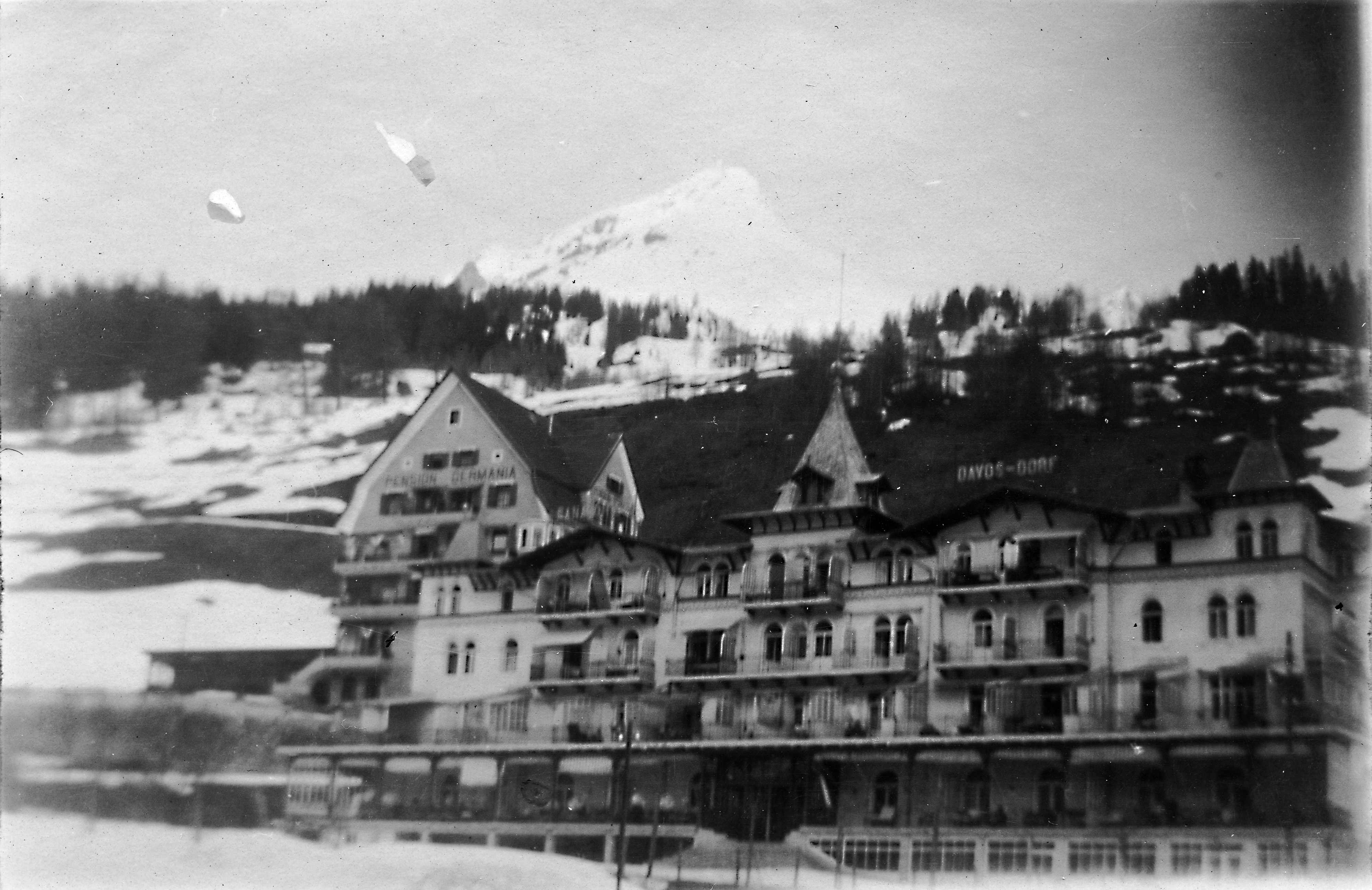 A black-and-white photograph of Sanatorium Davos-Dorf in snow: a long building with rows of balconies on every floor, pine trees and a mountain slope behind it.